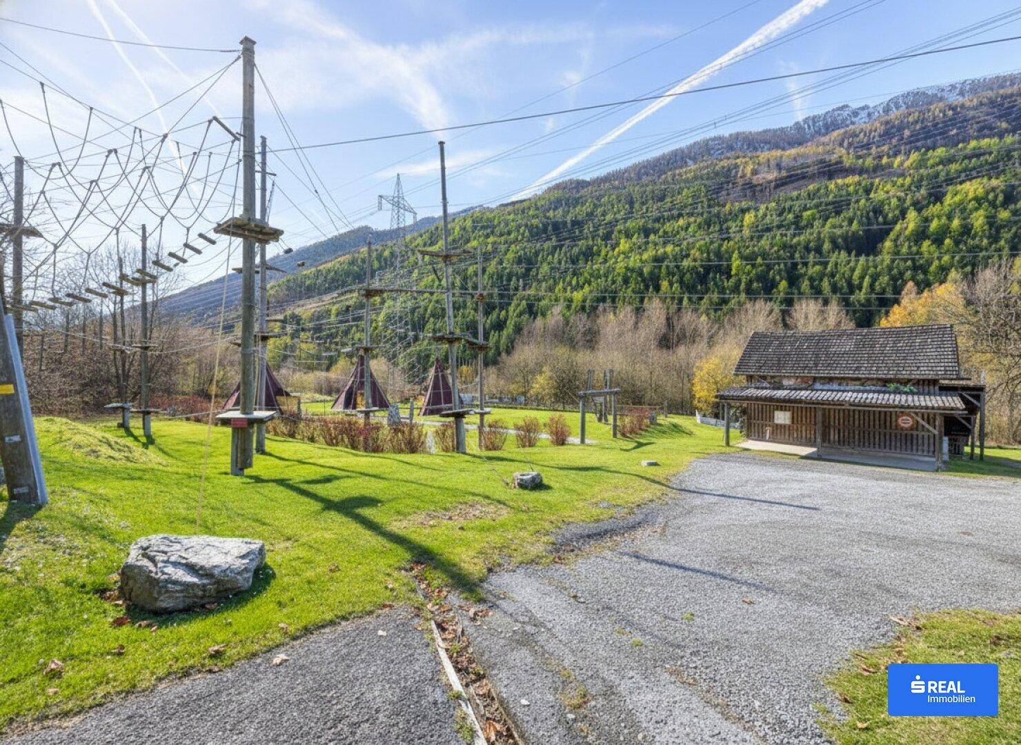 Outdoor-Erlebnispark mit Seilparcours und Holzhütte vor einer bewaldeten Bergkulisse.