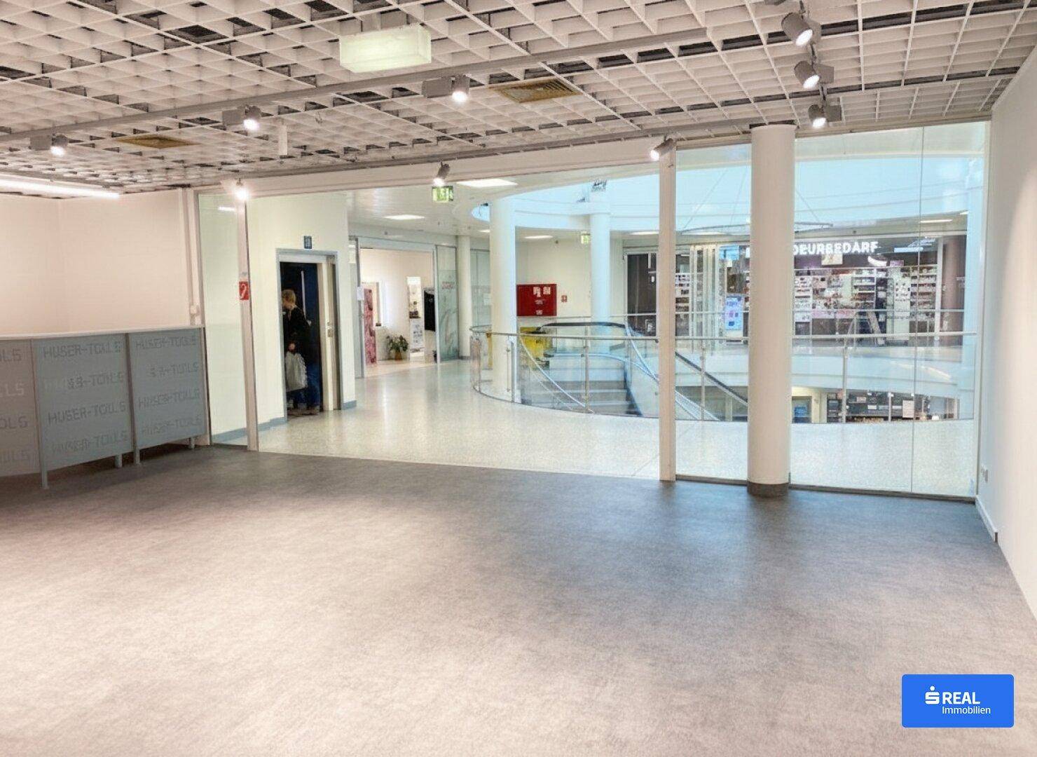 Breiter Flur in einem Gewerbegebäude mit Blick auf eine offene Ladenfläche und eine Wendeltreppe.