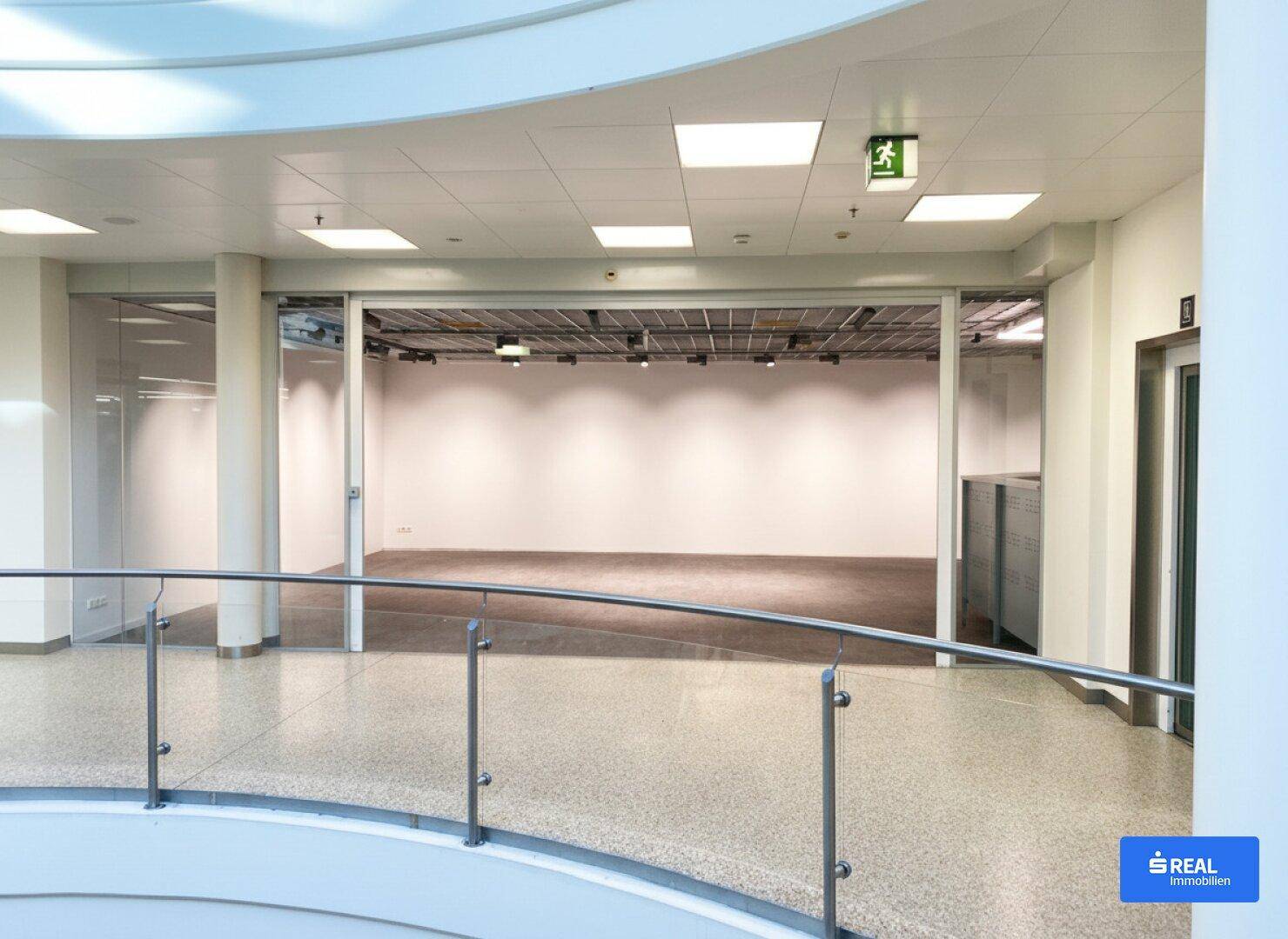Geräumiger, leerer Gewerberaum mit hohen Decken und großen Glasfronten, gesehen von einem oberen Flur.