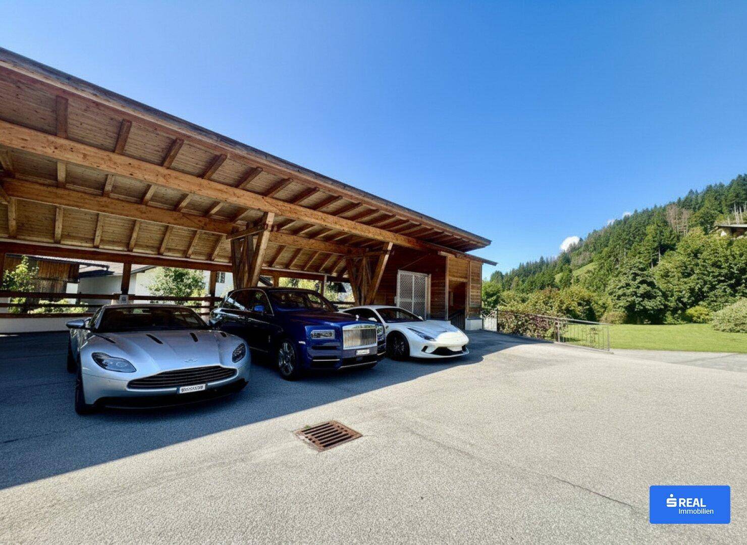 Großzügiger überdachter Parkplatz mit mehreren Luxusautos und Blick auf die bewaldeten Berge.
