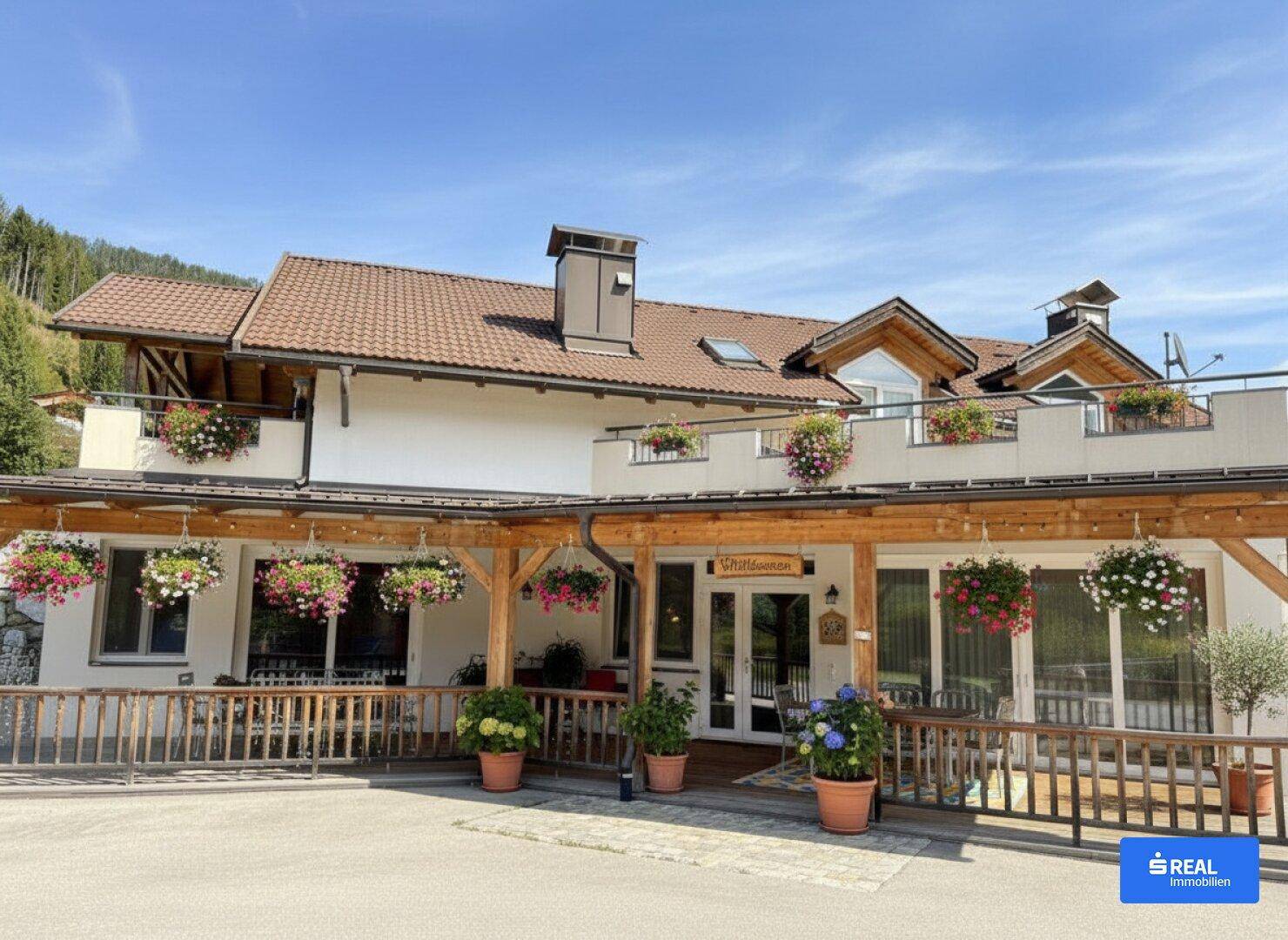 Traditionelles Gebäude mit überdachter Terrasse, vielen Blumen und Bergblick unter blauem Himmel.