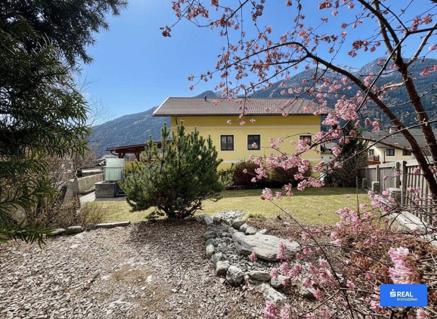 Gepflegter Gartenbereich mit blühenden Sträuchern und einem gelben Haus im Hintergrund, umgeben von Bergen.