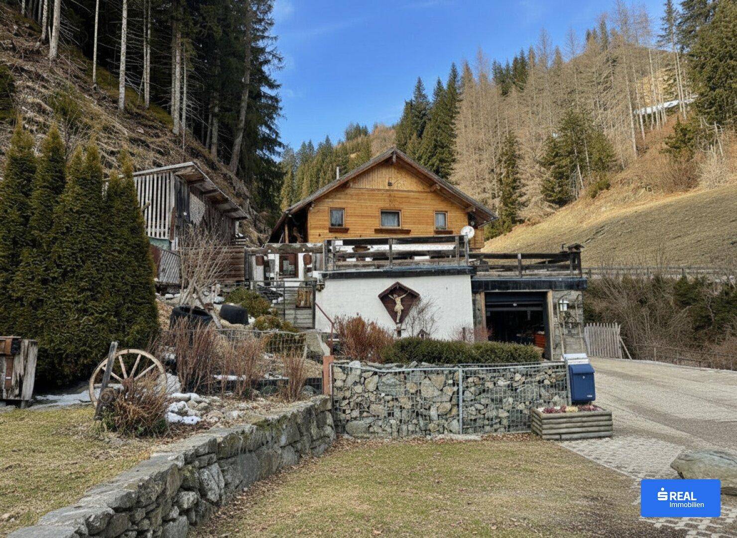 Rustikales Holzhaus in Hanglage mit Steingarten und Blick auf die umliegenden Wälder.