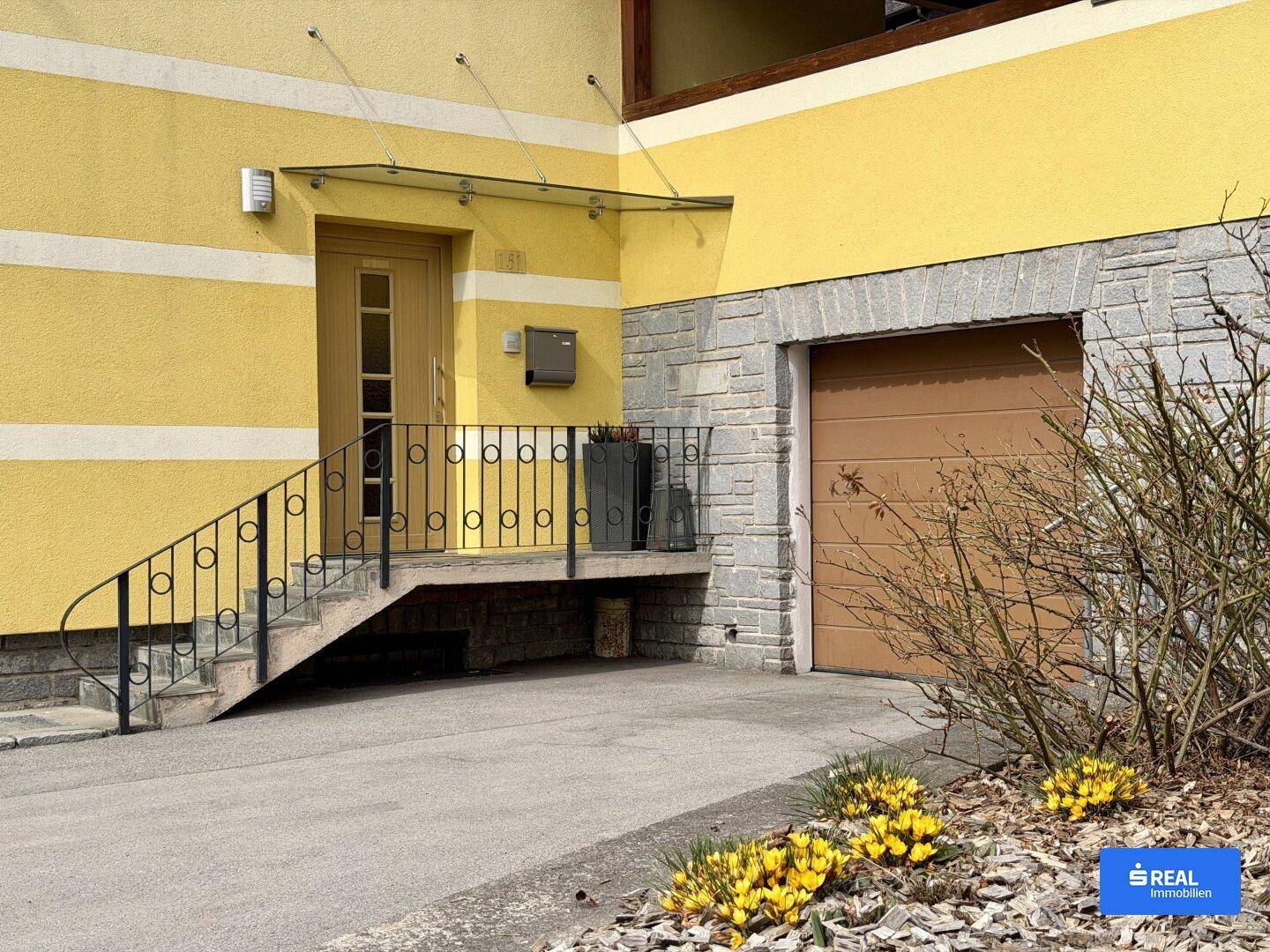 Hauseingang mit Treppe und Vordach, daneben eine Garage mit braunem Tor und Steinverkleidung.