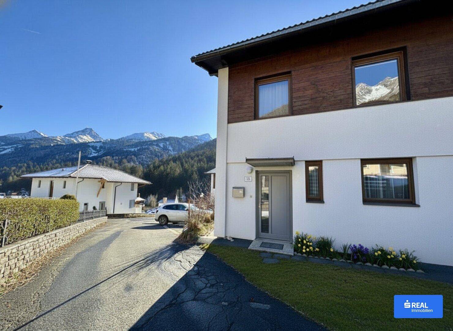 Seitenansicht des Hauses mit Einfahrt, Vorgarten und beeindruckendem Bergblick im Hintergrund.