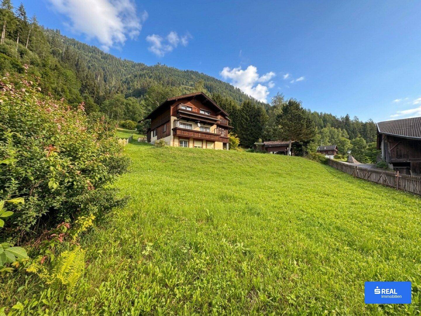 Traditionelles Holzhaus im Chalet-Stil auf einer grünen Wiese mit Bergblick unter blauem Himmel.