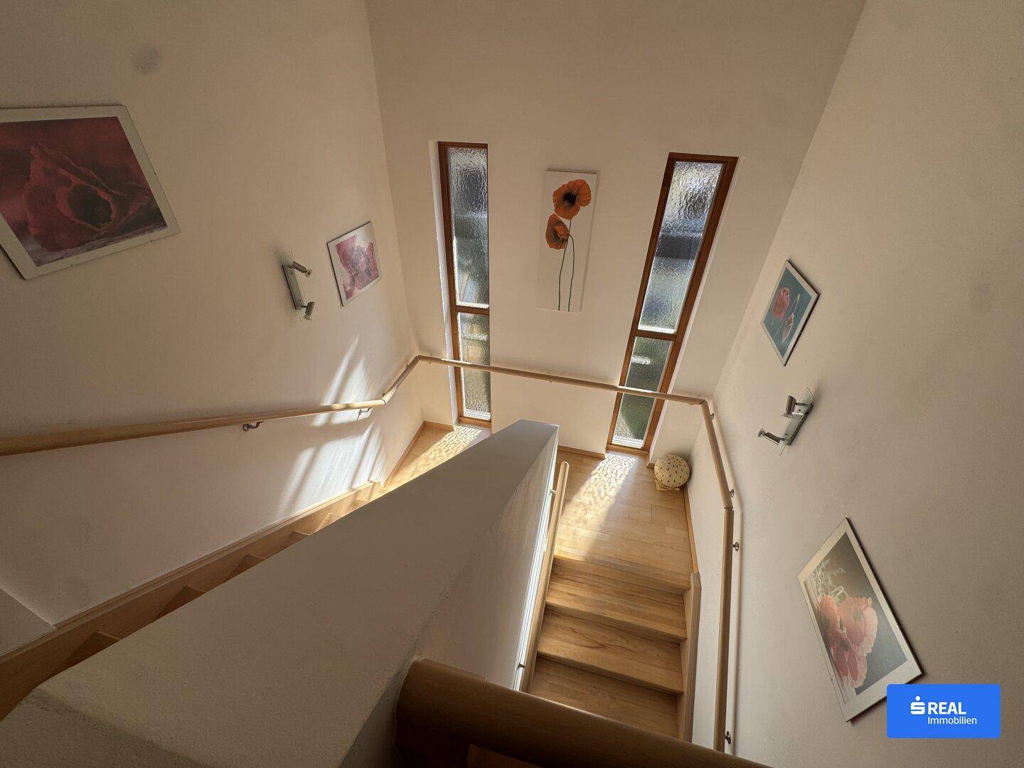Helles Treppenhaus mit Holzstufen und schmalen Fenstern, die viel Tageslicht hereinlassen.