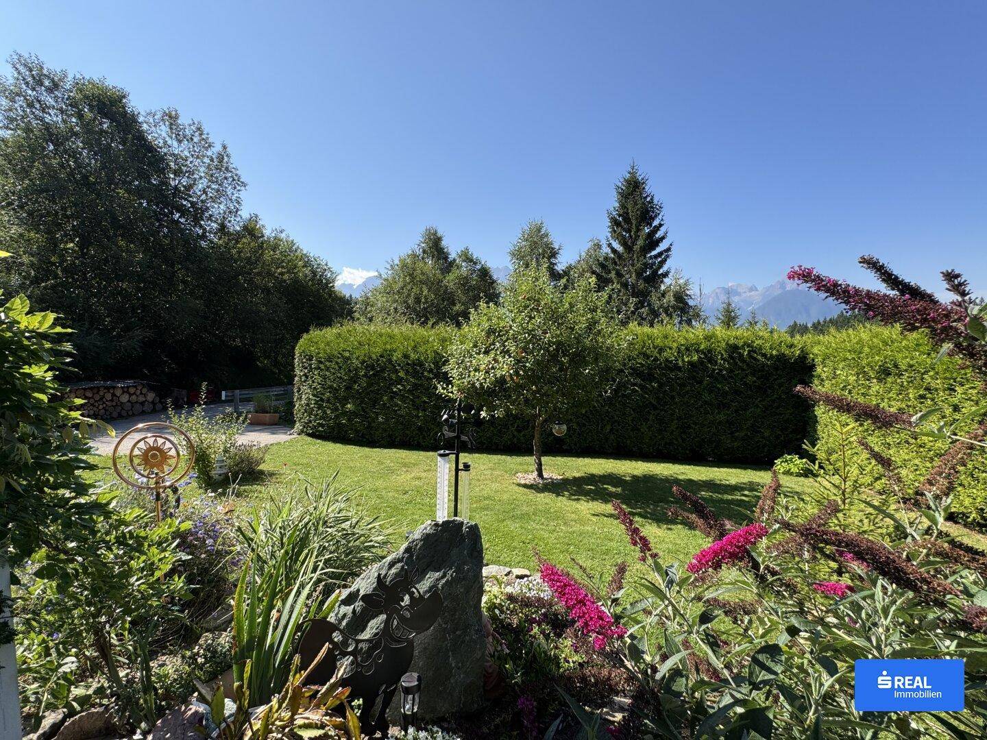 Blühender Garten mit Rasenfläche, hohen Hecken und einem weiten Blick in die Natur.