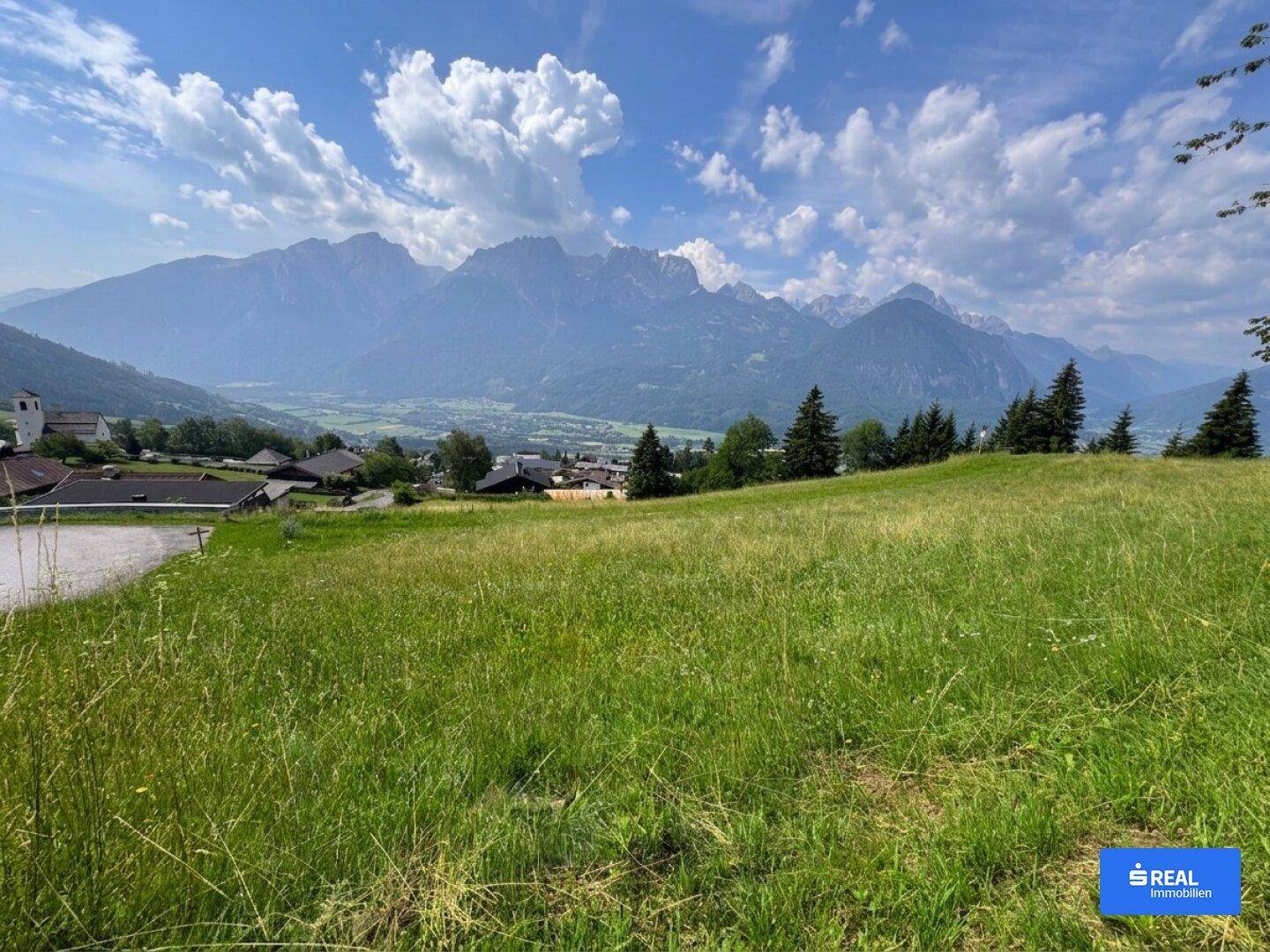 Sonnige Wiesenfläche mit Panoramablick auf die Alpen und eine kleine Ortschaft.