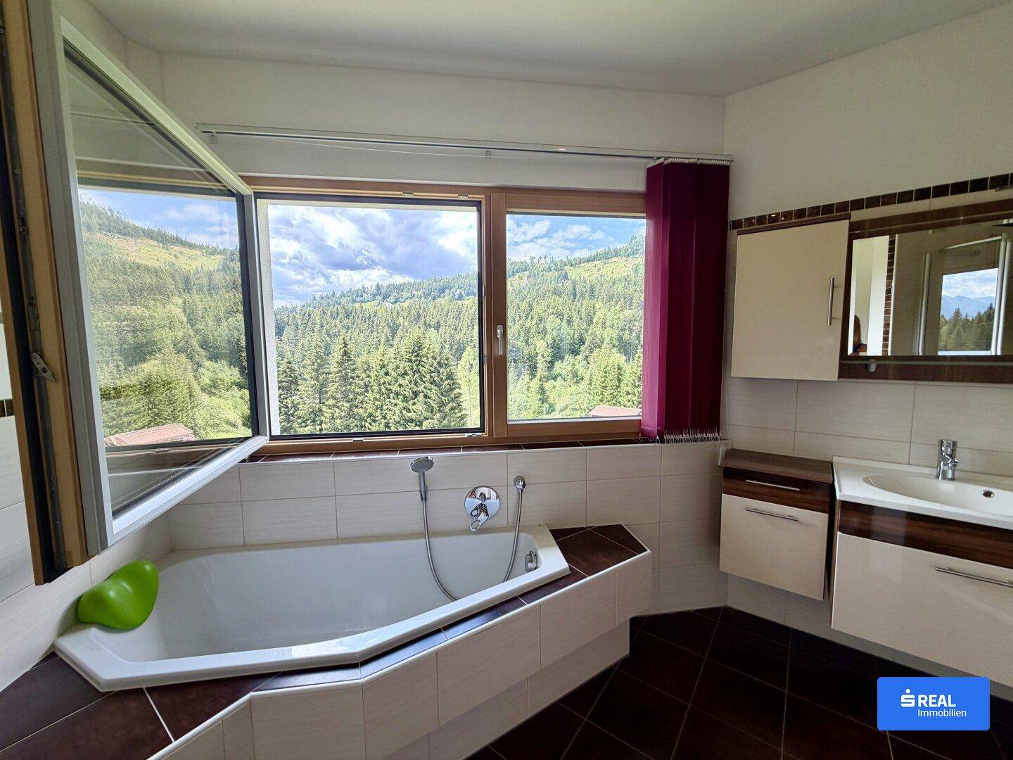 Helles Badezimmer mit Eckbadewanne, Einzelwaschbecken und Blick auf die bewaldete Landschaft.