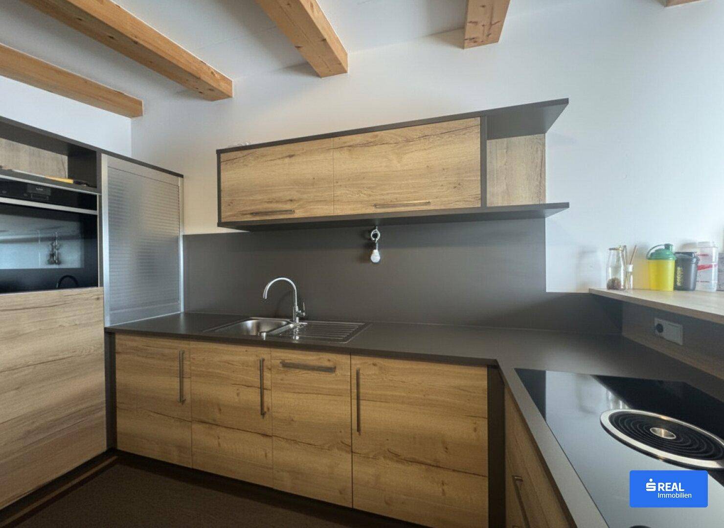 Moderne Küche mit Holzfronten, dunkler Arbeitsplatte und sichtbaren Deckenbalken, die eine gemütliche Atmosphäre schaffen.