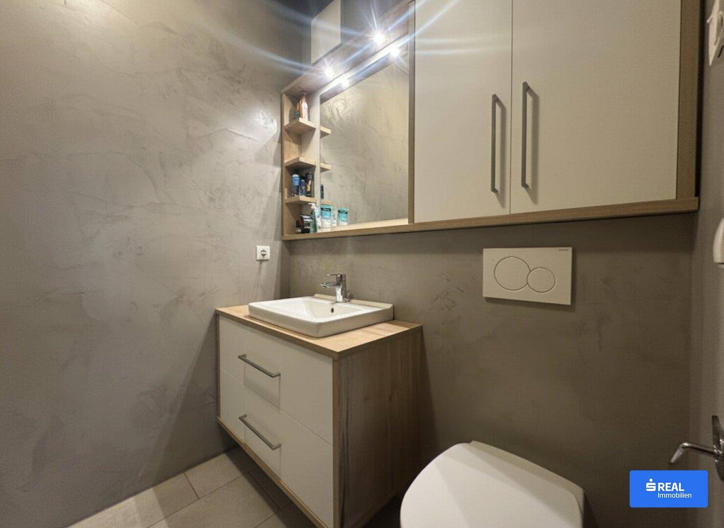 Modernes Badezimmer mit Waschbecken, Spiegelschrank und Hängeschränken, ergänzt durch eine Wandtoilette und strukturierte Wände.