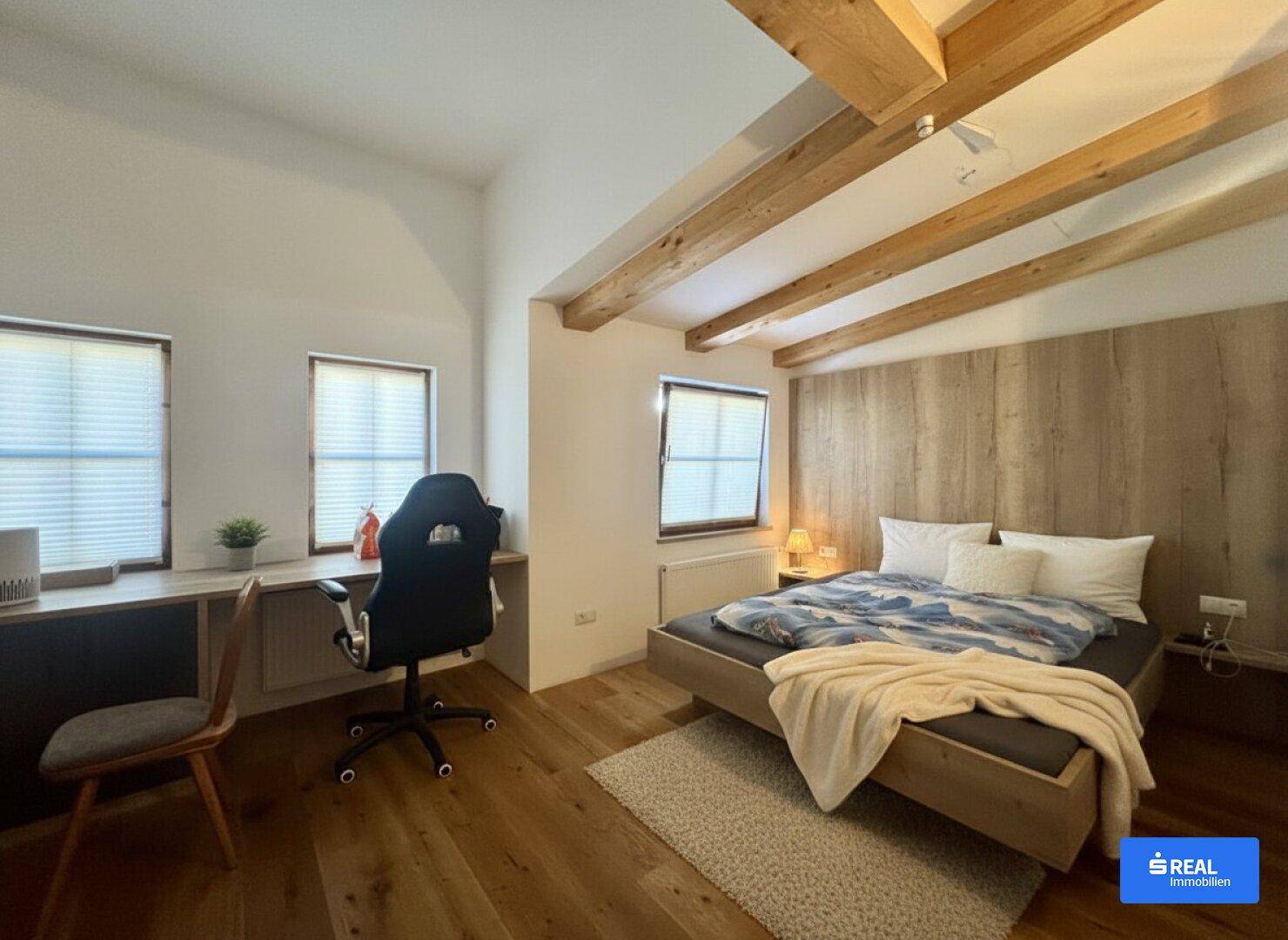 Gemütliches Schlafzimmer mit Holzbalkendecke, Holzboden und einem Arbeitsbereich am Fenster, der viel Tageslicht bietet.
