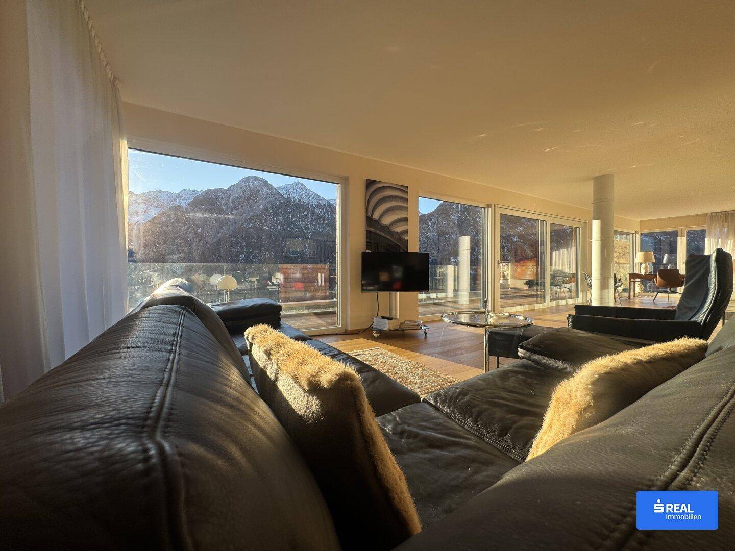 Helles Wohnzimmer mit großen Fenstern, die einen beeindruckenden Bergblick bieten. Moderne Einrichtung mit dunklem Ledersofa und Holzboden.