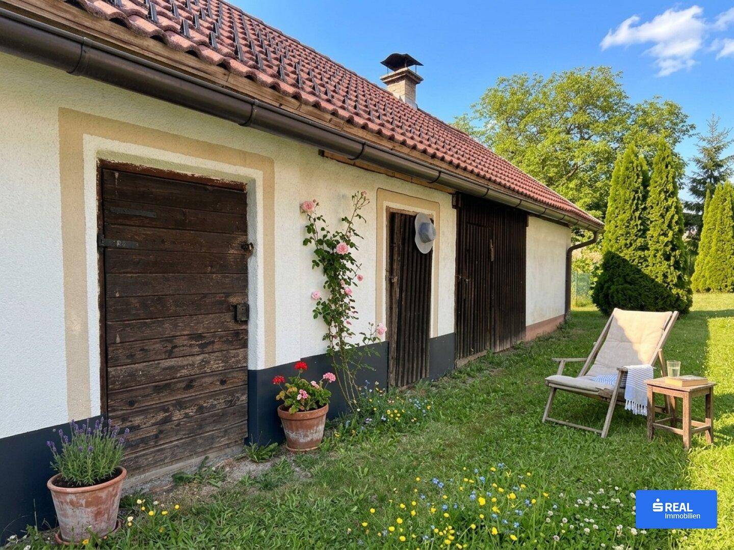 Seitlicher Bereich des Hauses mit rustikalen Holztüren, Blumen und einem Liegestuhl im Grünen.