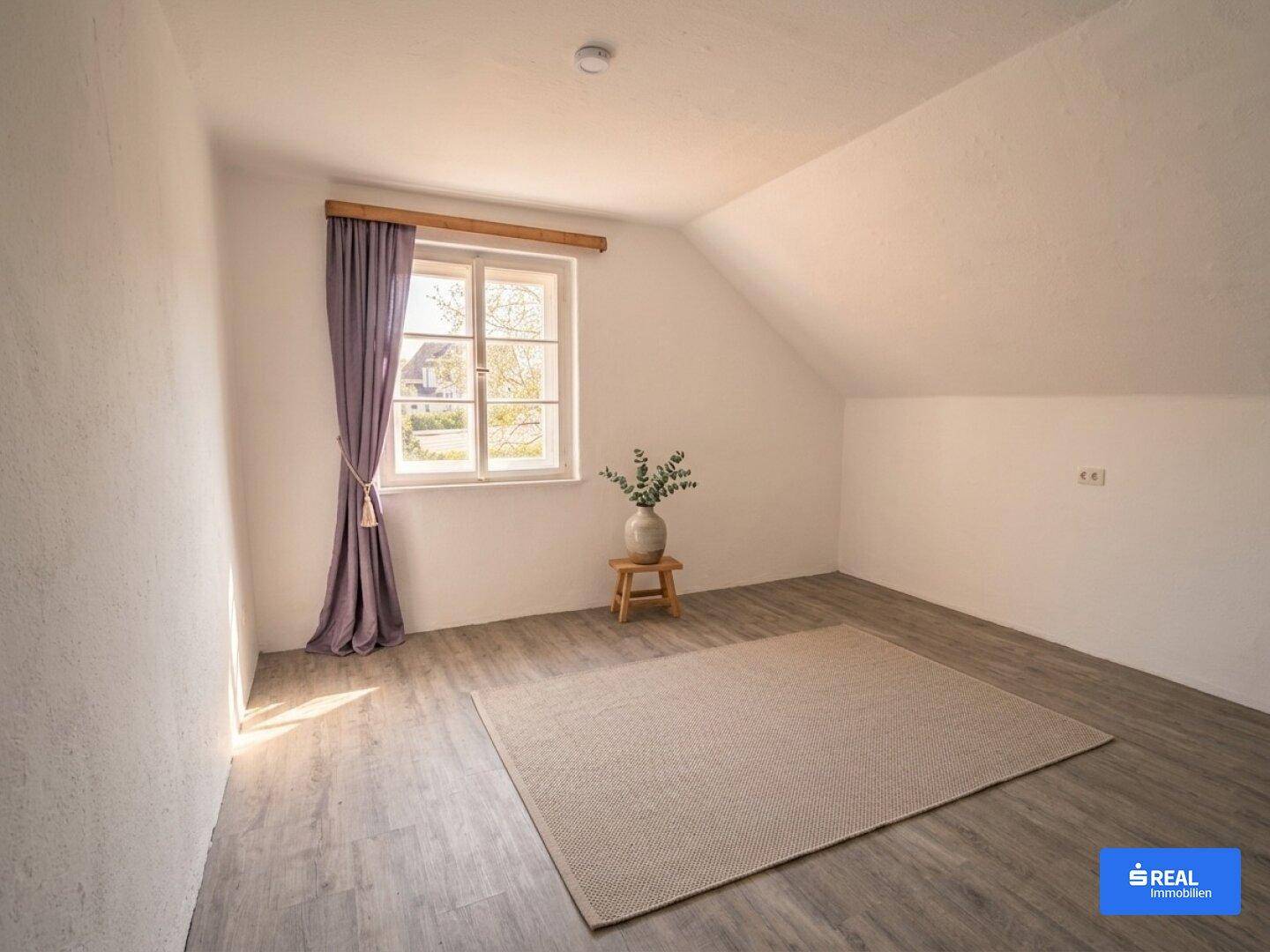 Heller Raum im Dachgeschoss mit schrägen Wänden, Fenster und modernem Bodenbelag.