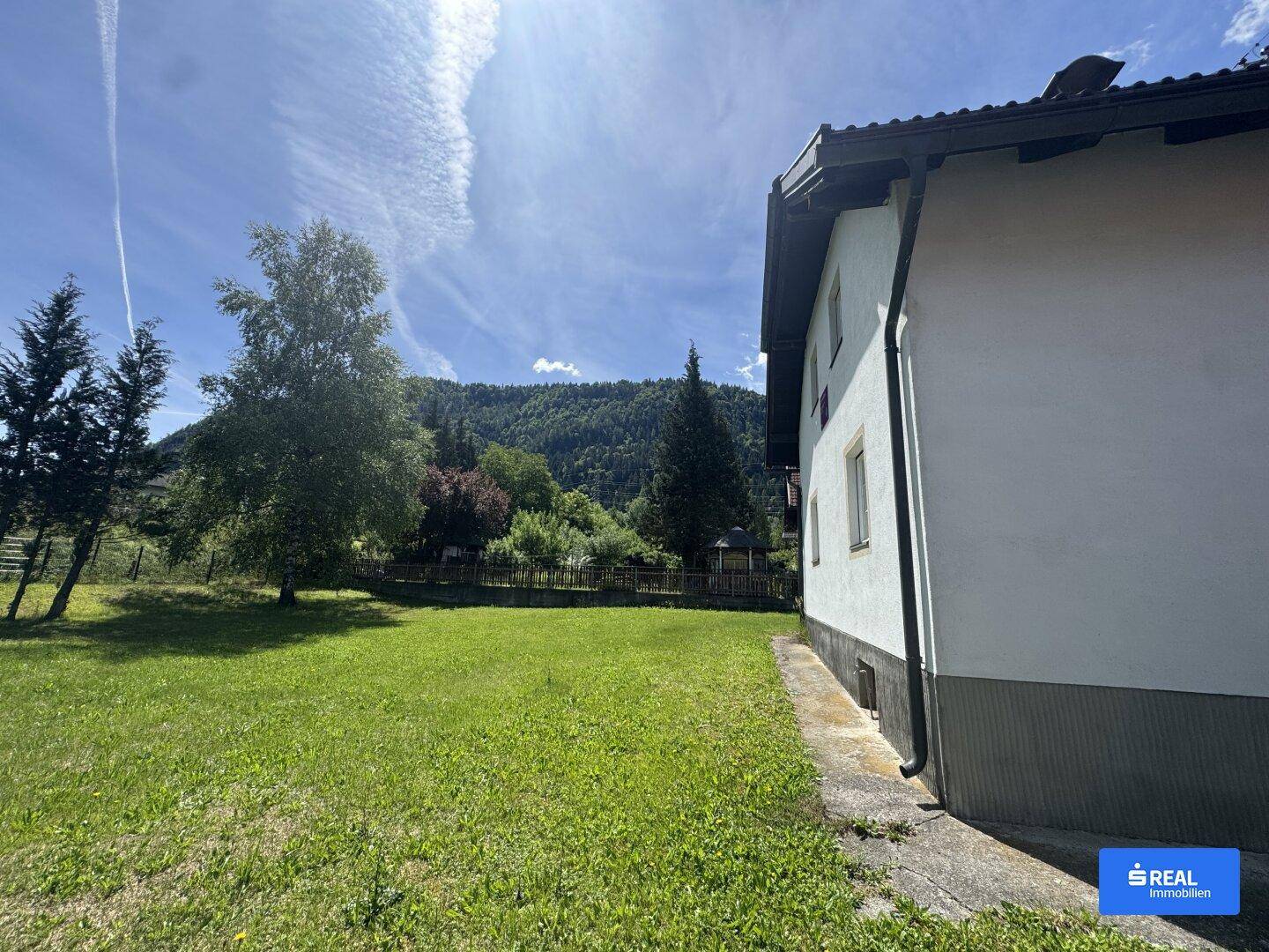 Sonniger Außenbereich des Hauses mit weitem Rasen und Blick auf die bewaldeten Berge.