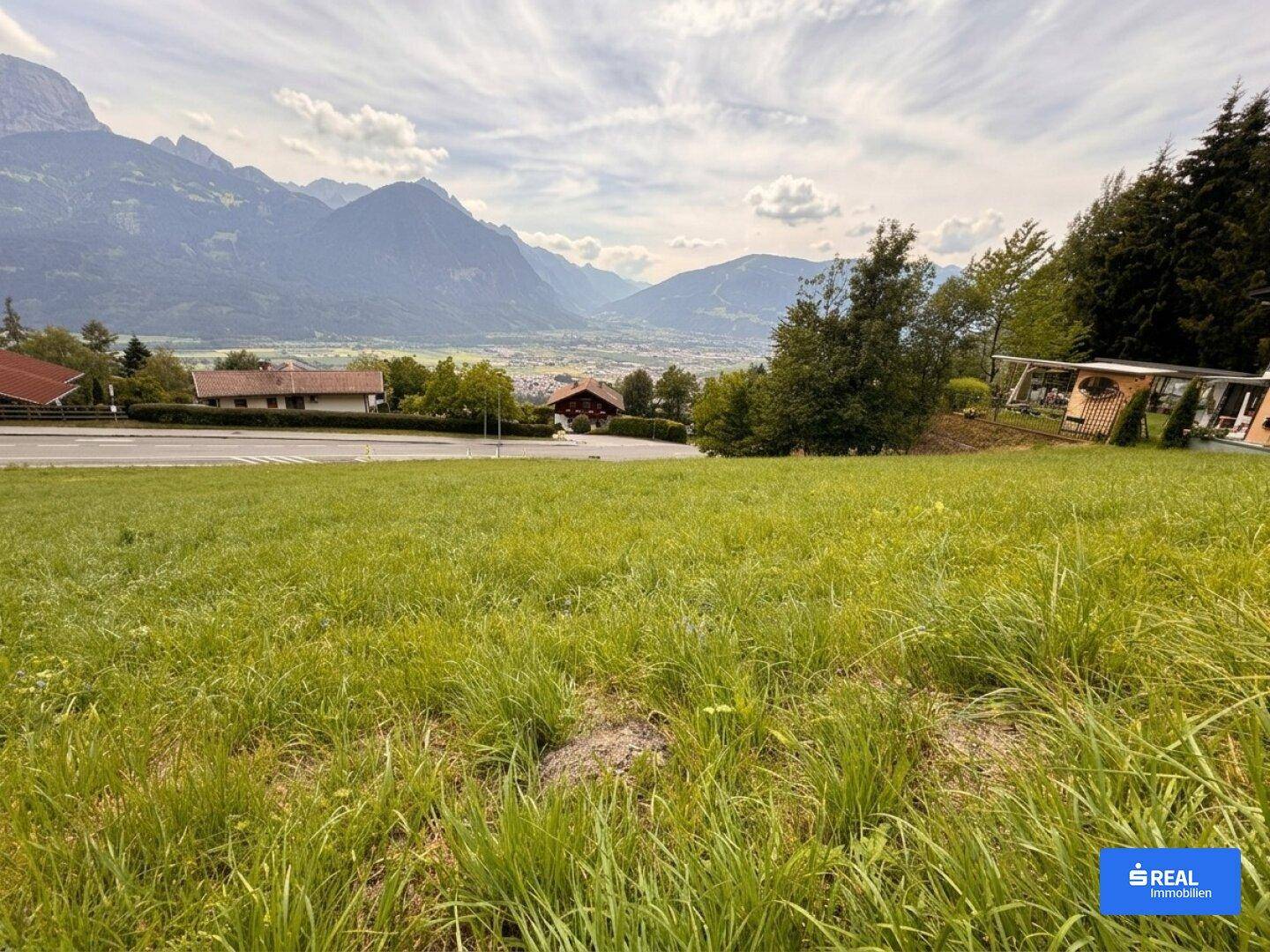 Große grüne Wiese mit Blick auf die umliegende Berglandschaft und einige Wohnhäuser.