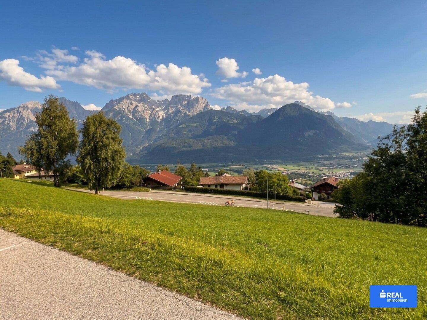 Weitläufige grüne Wiese mit Blick auf eine beeindruckende Bergkette und ein sonniges Tal.