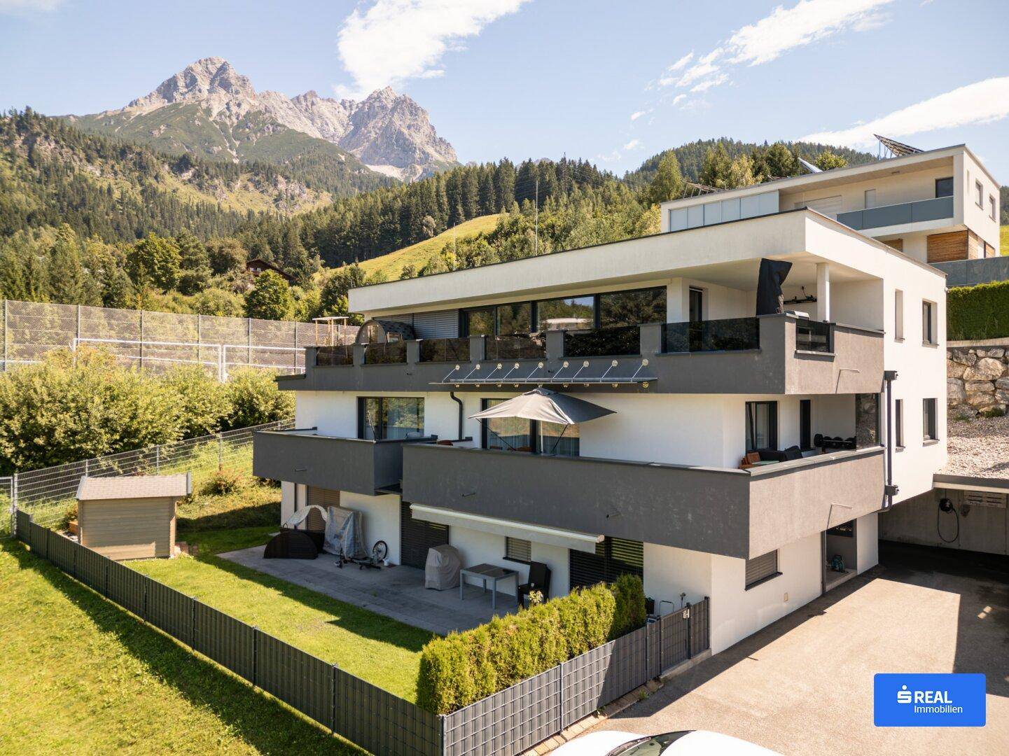 Ansicht des modernen Apartmenthauses mit mehreren Balkonen und einem kleinen Gartenbereich vor alpiner Kulisse.