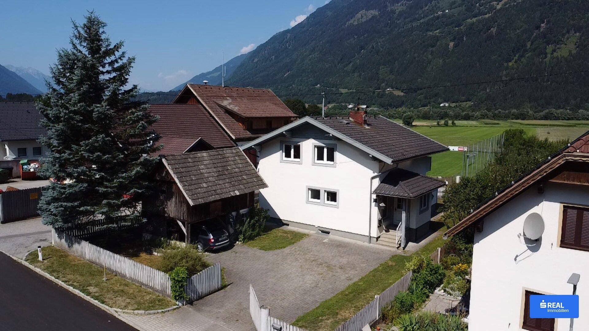 Rückansicht des Hauses mit Carport und Auffahrt, umgeben von grüner Landschaft und Bergen.