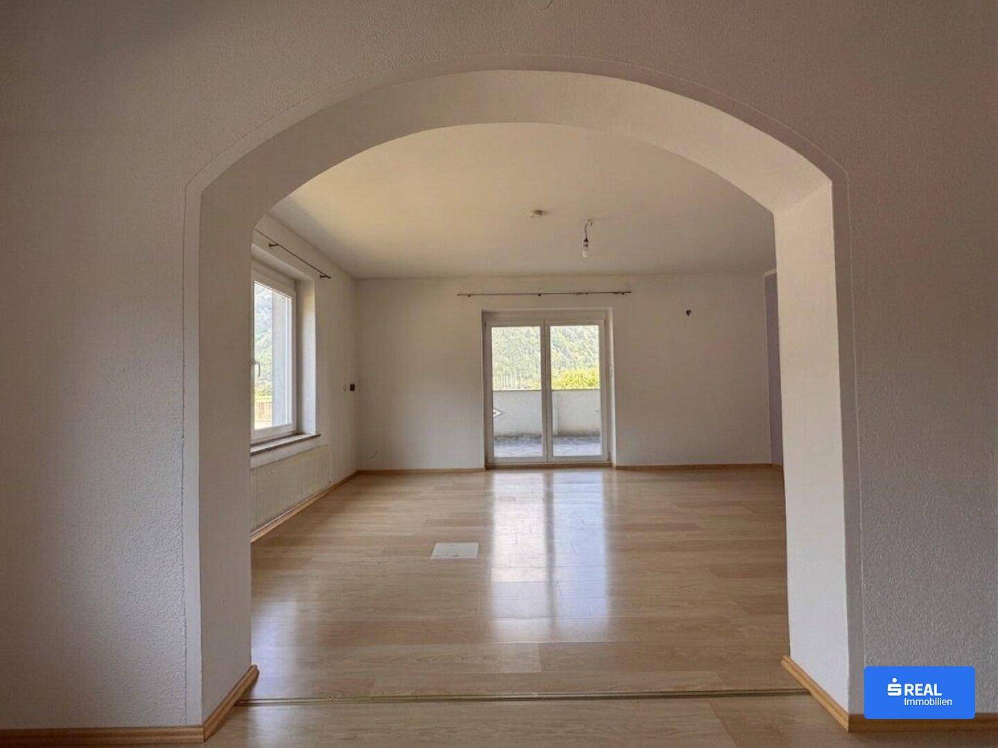 Heller, leerer Wohnraum mit Holzboden, Bogenöffnung und Zugang zu einem Balkon mit Bergblick.