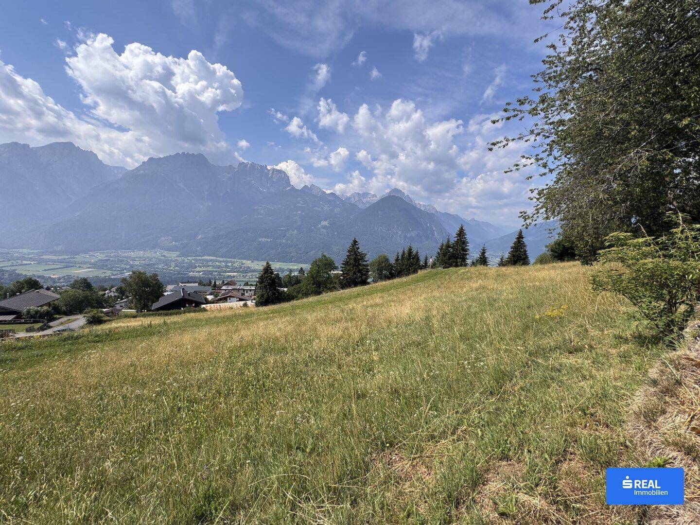 Panoramablick über grüne Wiesen und ein Tal mit Häusern, umgeben von einer beeindruckenden Bergkulisse.