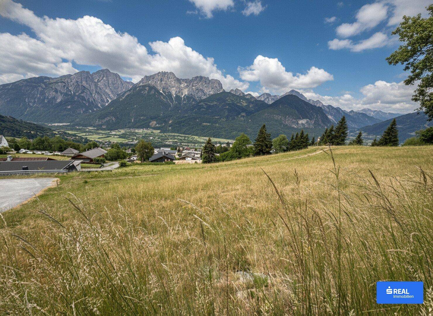 Malerische Landschaft mit weiten Feldern, einem Dorf im Tal und hohen Bergen unter einem strahlend blauen Himmel.