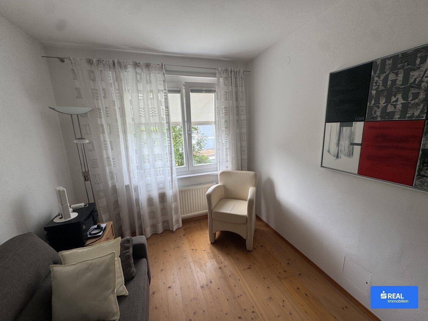 Helles Zimmer mit Holzboden, einem bequemen Sessel und einem Fenster mit Tageslicht.