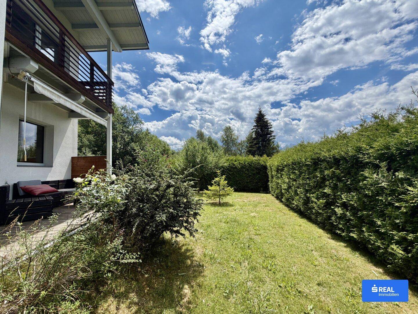 Gepflegter Gartenbereich mit Rasenfläche, hoher Hecke und einer gemütlichen Terrasse unter einem Balkon.
