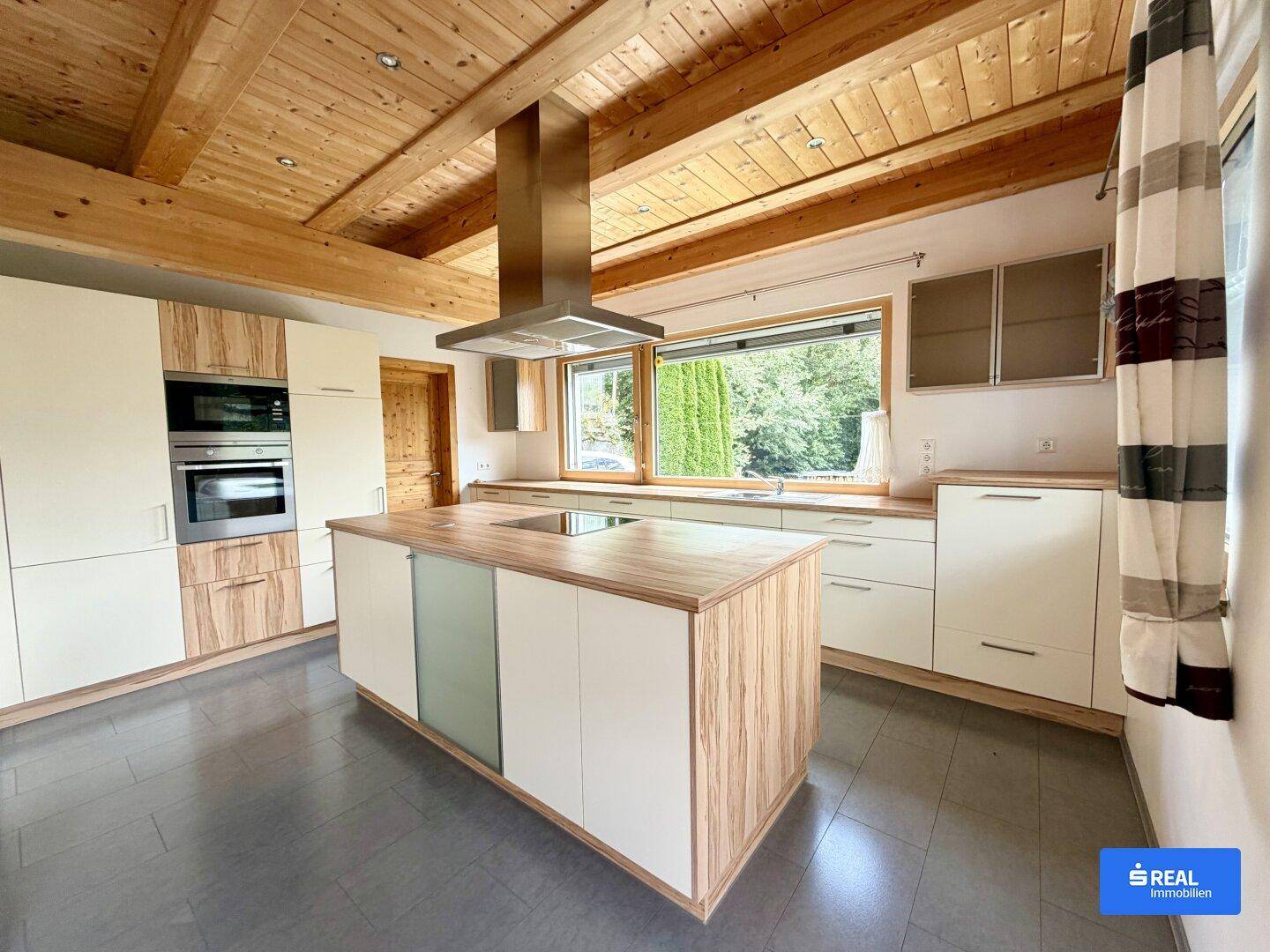 Moderne Küche mit Holzbalkendecke, Kücheninsel und integrierten Geräten, hell durch großes Fenster.