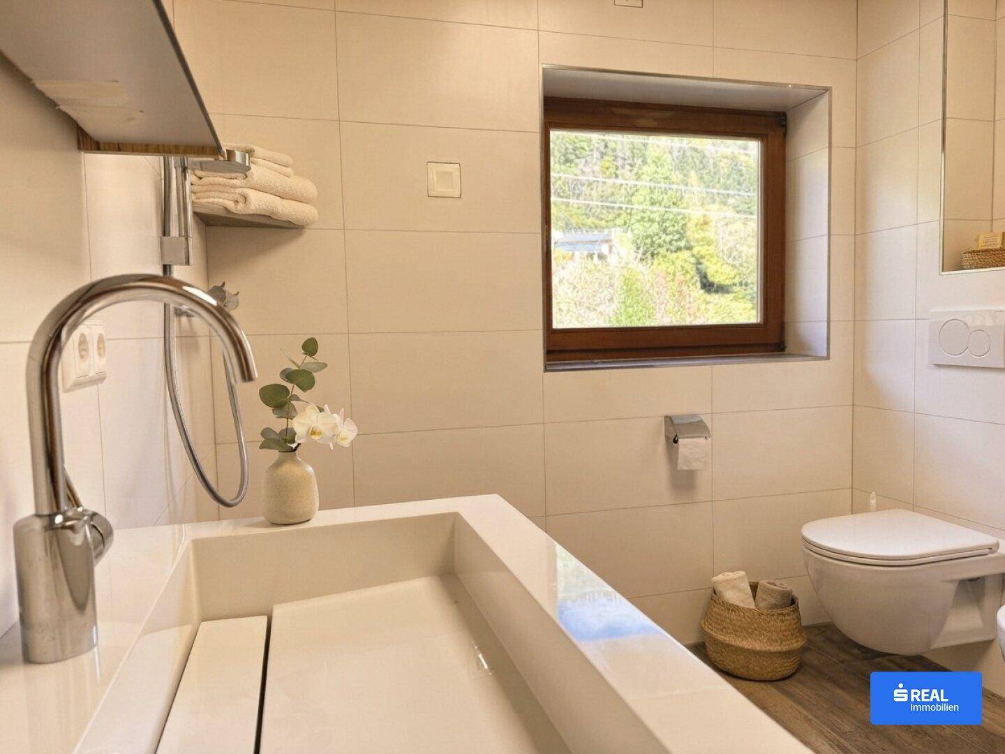 Helles Badezimmer mit Wand-WC, modernem Waschbecken und Fenster mit Blick ins Grüne.