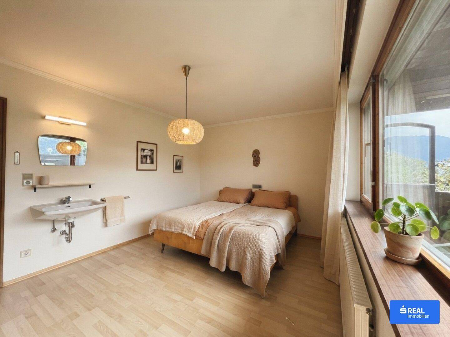 Helles Schlafzimmer mit Doppelbett, Holzboden, Waschbecken und großem Fenster mit Bergblick.
