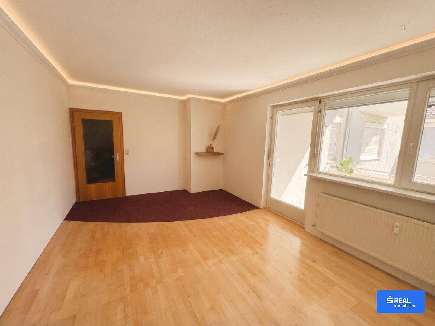 Geräumiges Wohnzimmer mit hellem Parkettboden, großen Fenstern und Zugang zu einem Außenbereich.