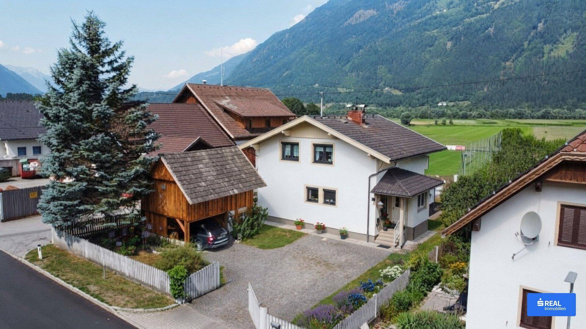 Haus mit Carport und Garten in ländlicher Umgebung mit Bergblick und viel Grün.