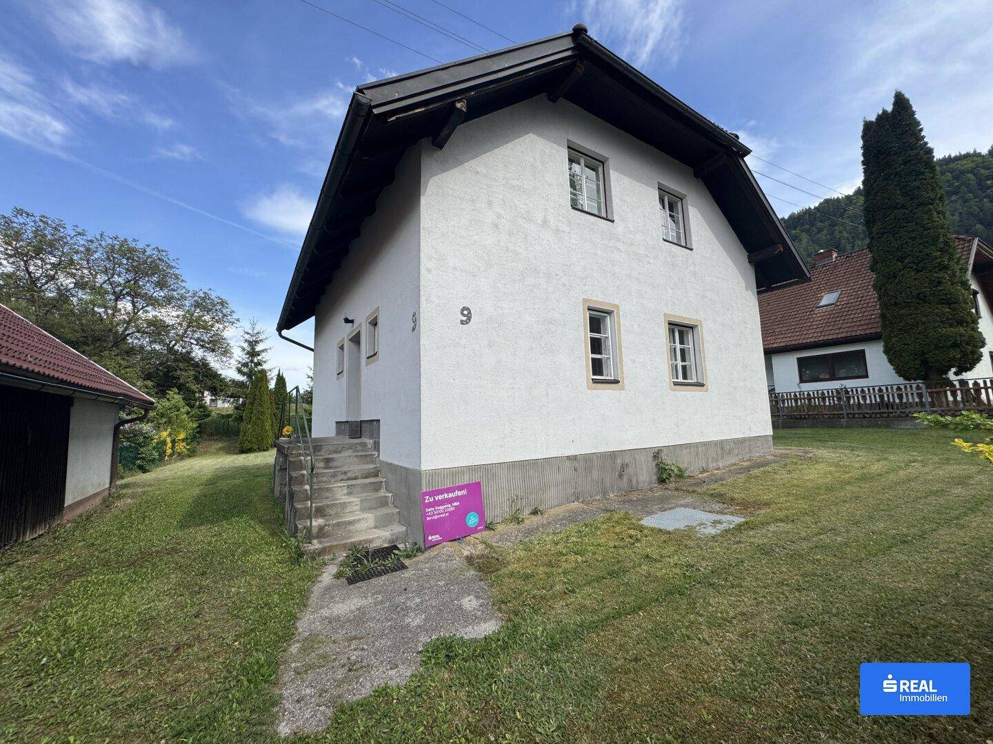 Weißes Einfamilienhaus mit dunklem Satteldach und gepflegtem Vorgarten unter blauem Himmel.