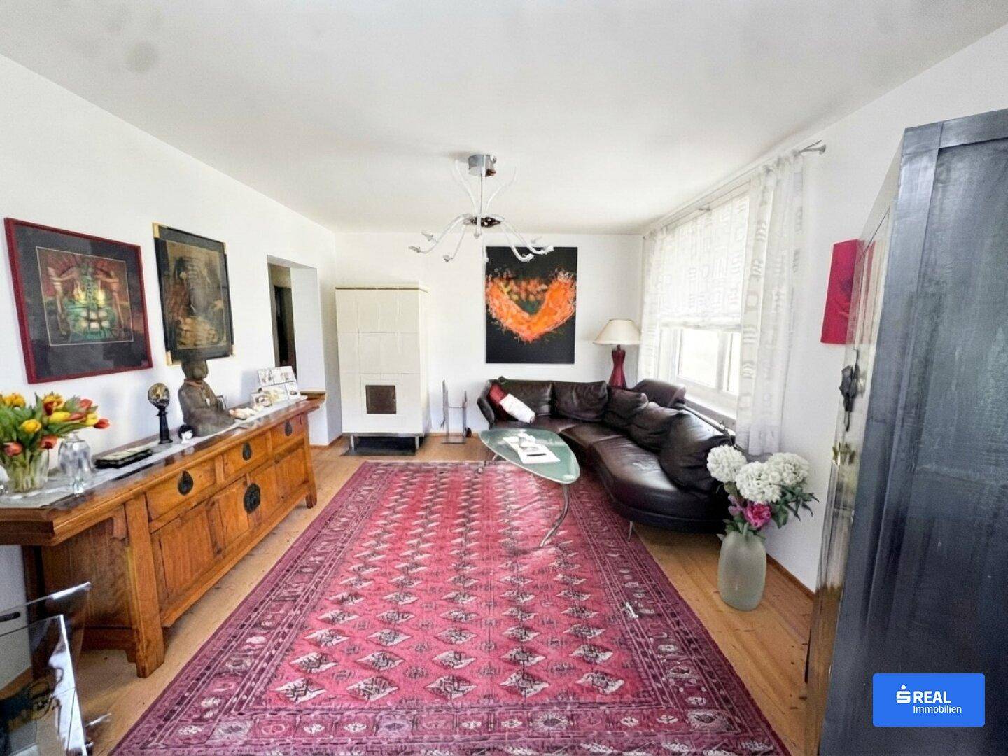 Geräumiges Wohnzimmer mit dunklem Ledersofa, Holzboden, Kachelofen und einem großen orientalischen Teppich.