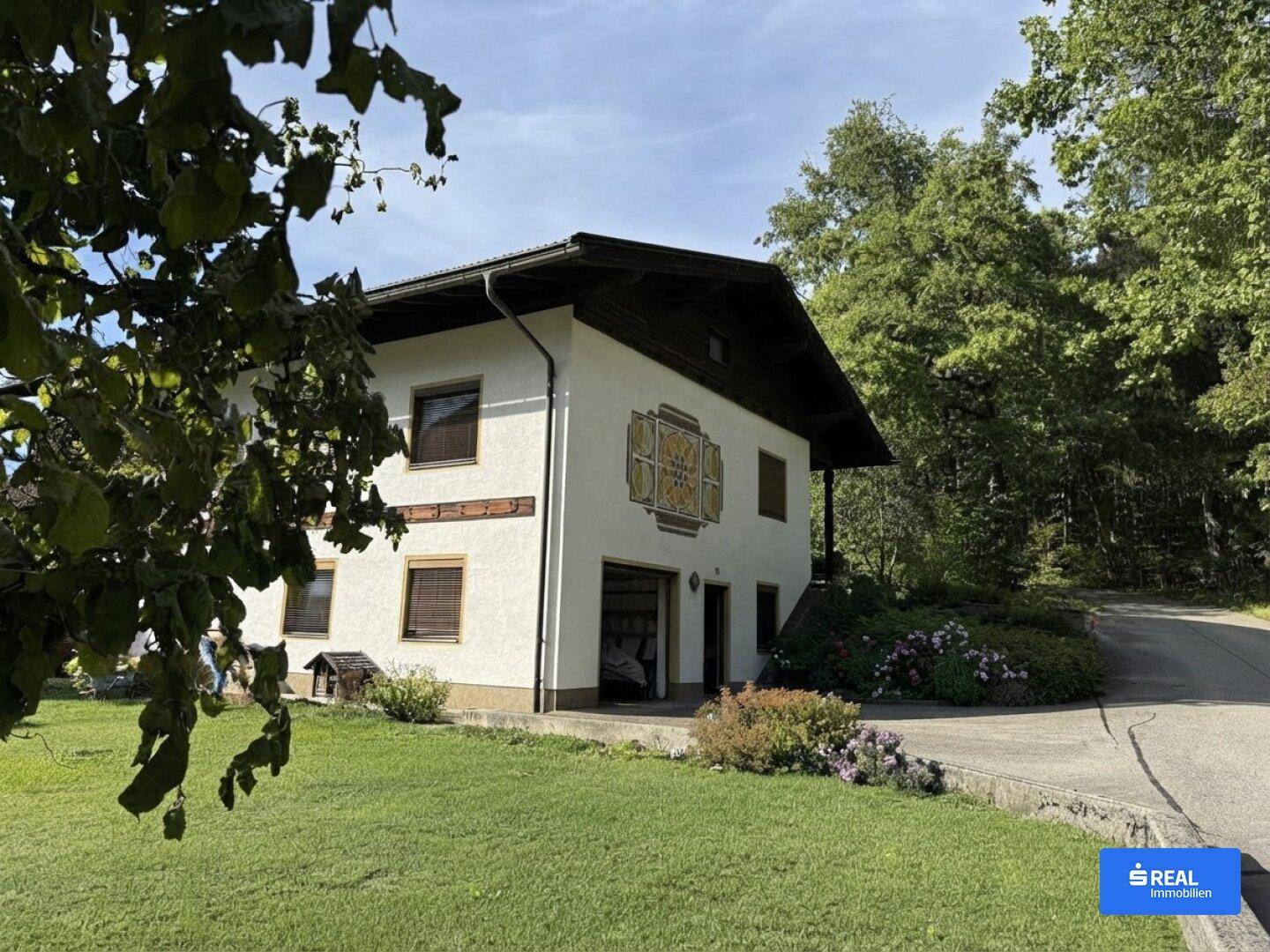 Ansicht des Hauses mit Garage, dekorativer Fassadenmalerei und Zufahrt, umgeben von Grünflächen.