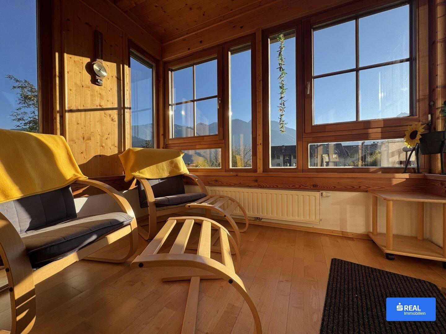 Lichtdurchfluteter Wintergarten mit Holzvertäfelung, bequemen Liegestühlen und Panoramablick auf die Berge.