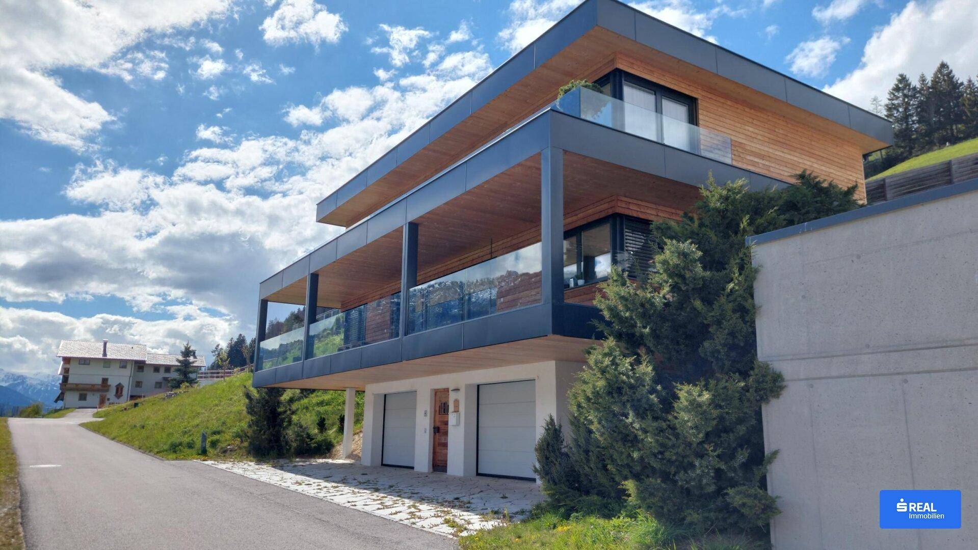 Moderne Architektur mit Holzverkleidung und großen Balkonen, eingebettet in eine grüne Hanglage unter blauem Himmel.