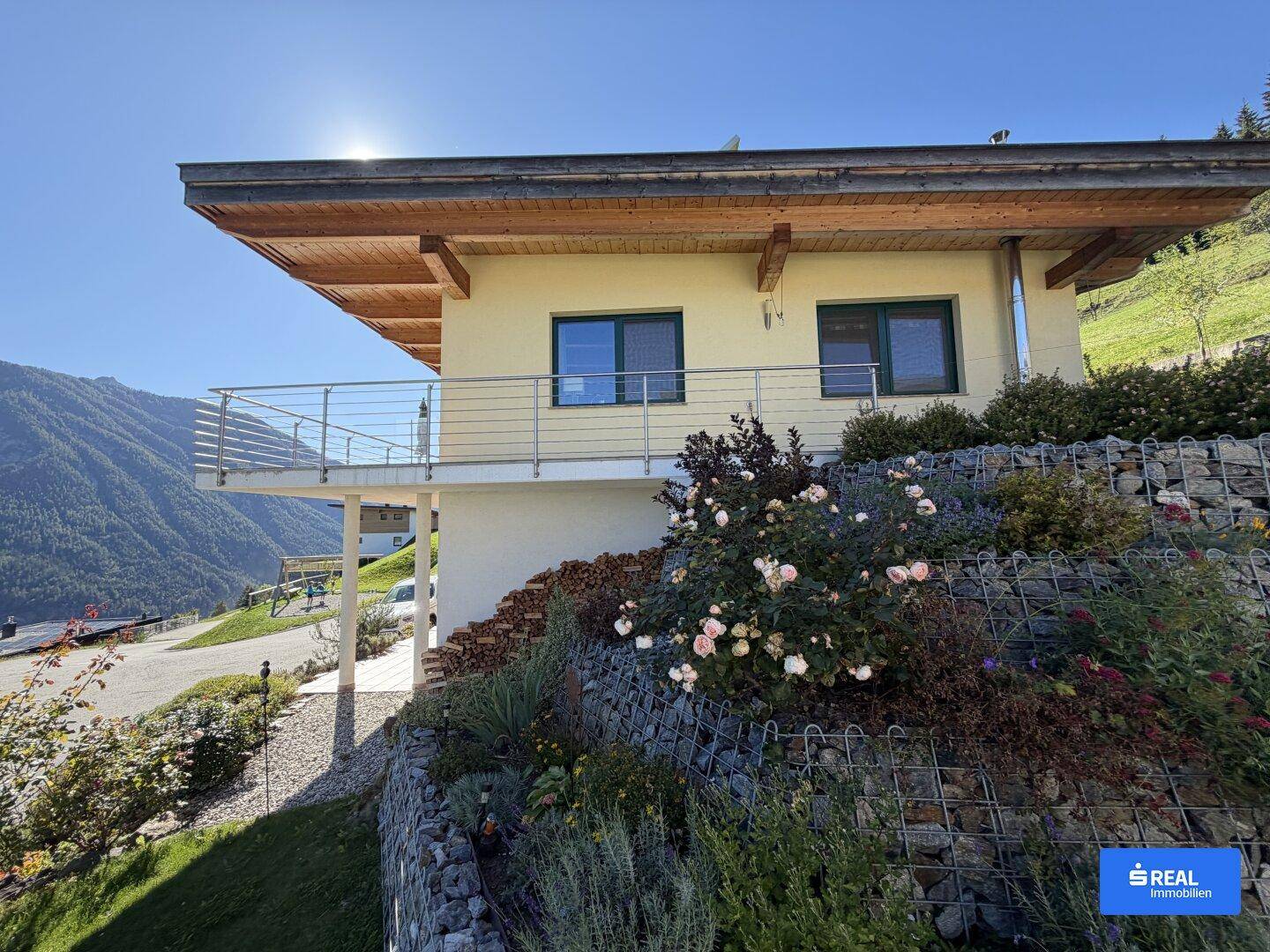 Hausfassade mit Balkon, Gabionenmauer und blühenden Rosen vor einer malerischen Berglandschaft.