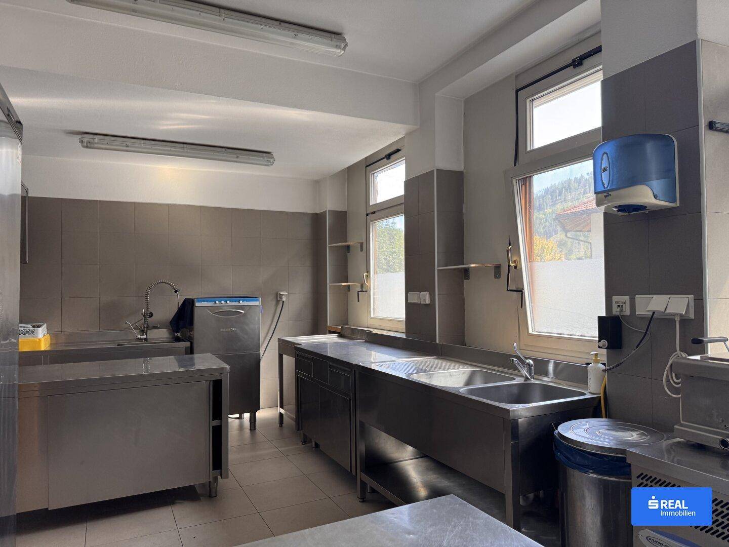 Geräumige Gewerbeküche mit mehreren Edelstahlspülbecken, Arbeitsflächen und Fenstern für ausreichend Licht.