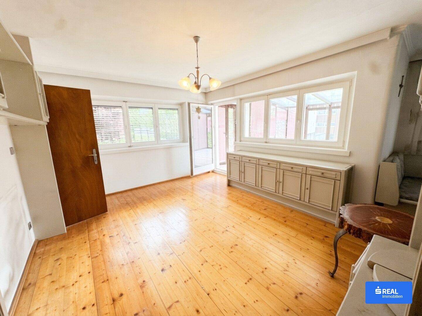 Heller Wohn- und Essbereich mit Holzdielenboden, großen Fenstern und einem Sideboard.