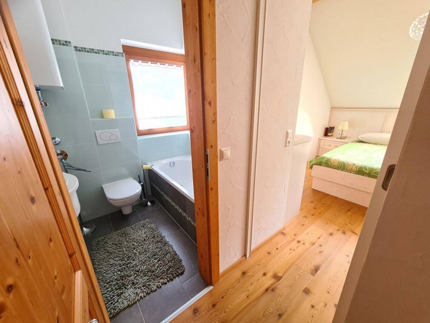 Blick vom Flur in ein Badezimmer mit Badewanne und ein angrenzendes Schlafzimmer mit Holzboden.