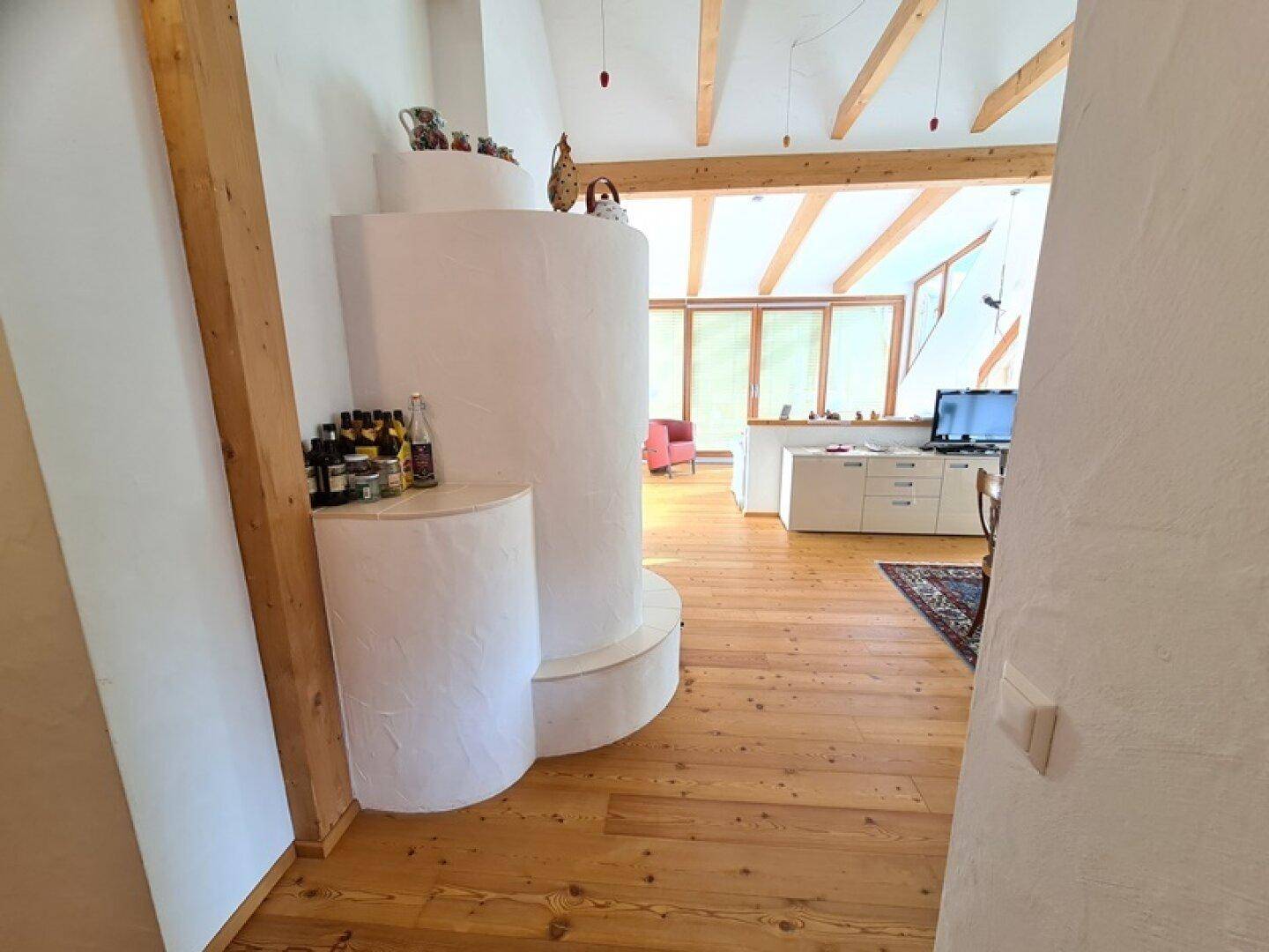 Offener Wohnbereich mit Kachelofen, Holzboden und Blick auf einen Ess- oder Küchenbereich.