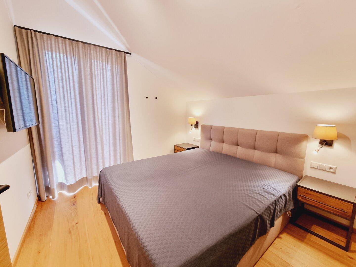 Geräumiges Schlafzimmer mit Doppelbett, Fernseher und großem Fenster mit Vorhängen.