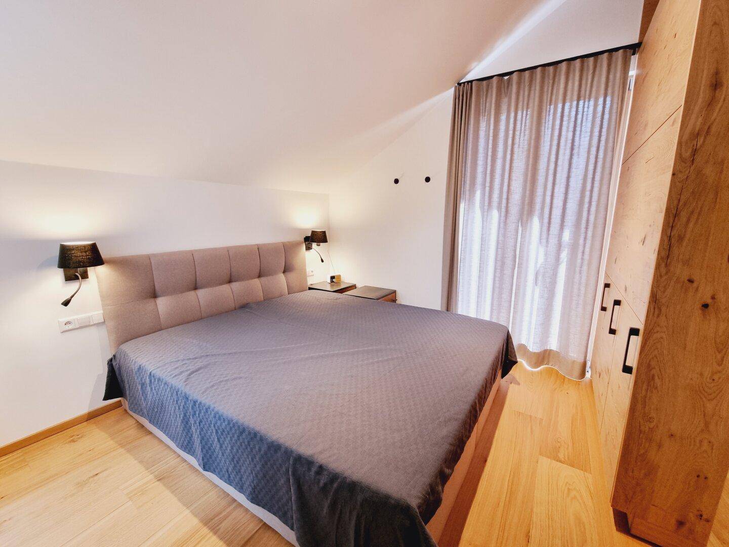 Schlafzimmer mit Doppelbett, Holzschrank und großem Fenster, das viel Licht hereinlässt.