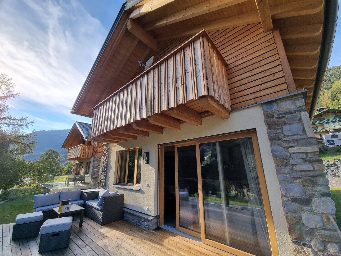 Holzterrasse mit Sitzgelegenheiten und Balkon darüber, umgeben von grüner Berglandschaft.