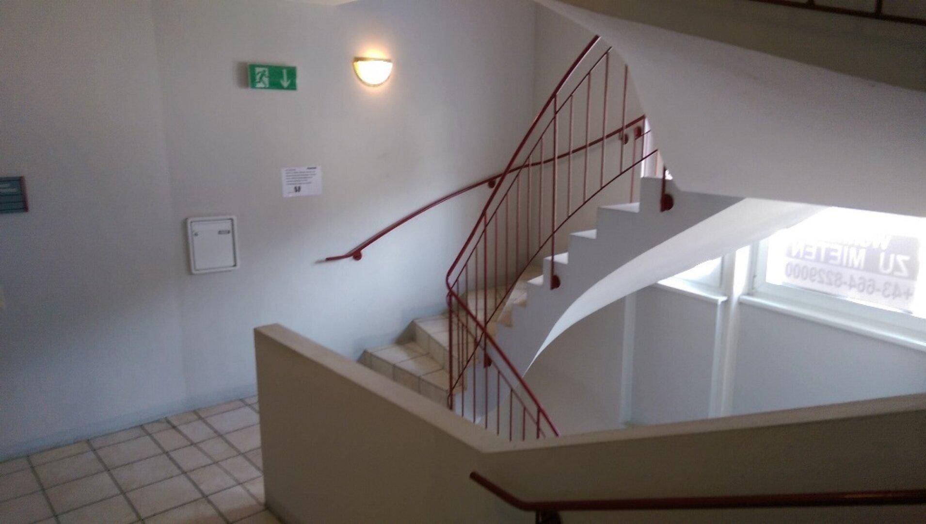 Helles Treppenhaus mit rotem Geländer, Fliesenboden und natürlichem Lichteinfall durch ein Fenster.