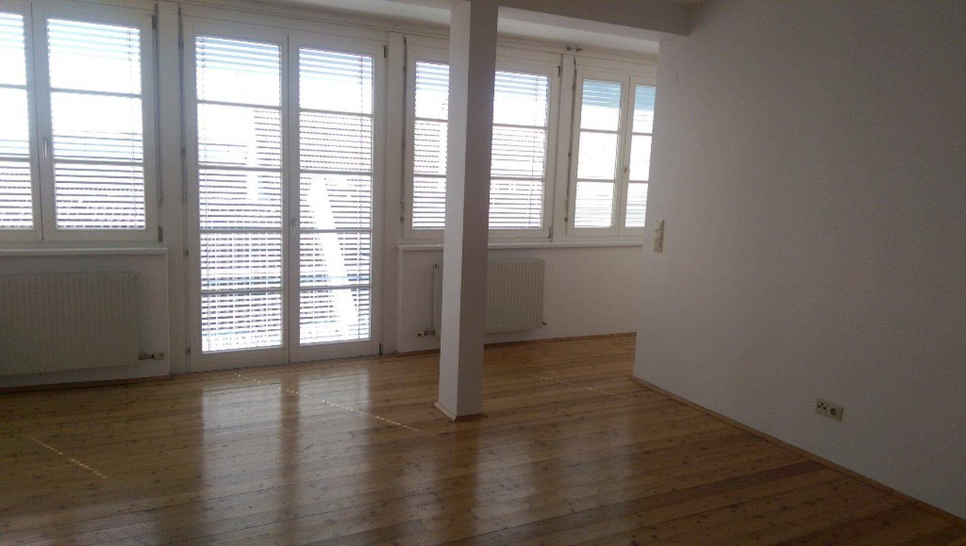 Heller Wohnbereich mit großen Fenstern, Holzfußboden und einem tragenden Pfeiler.